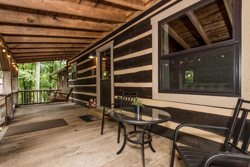 Covered wooden porch with a glass-top table, black metal chairs, a swinging bench, and string lights.