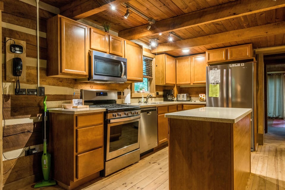 Kitchen with honey-toned wood cabinets, stainless steel appliances, and a center island under exposed log beams.
