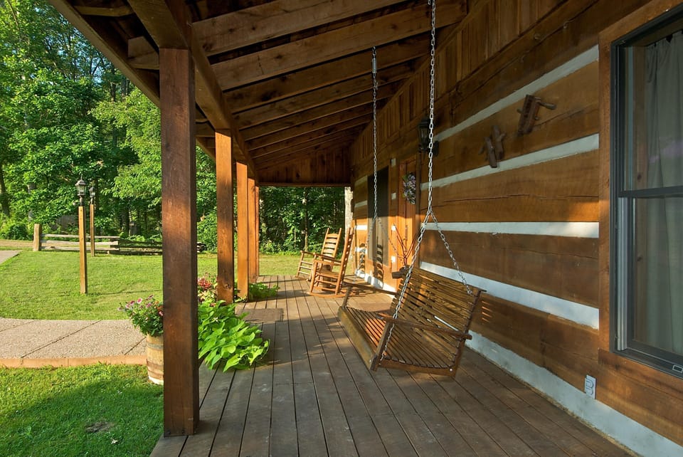 Covered porch with wooden swing, rocking chairs, and log cabin siding overlooking a green lawn and trees.