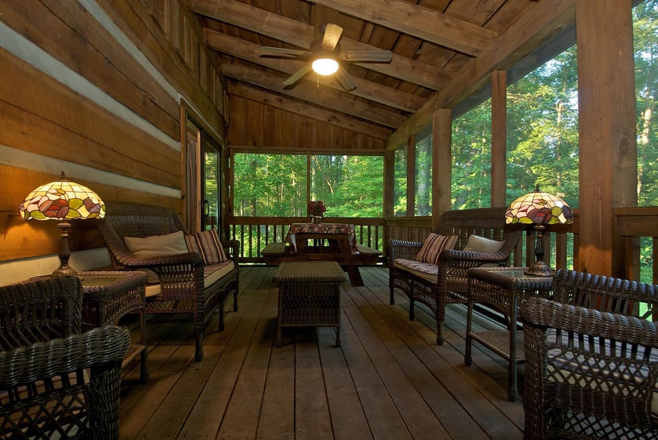 Screened porch with wicker seating, two Tiffany-style lamps, a ceiling fan, and a picnic table overlooking wooded views.