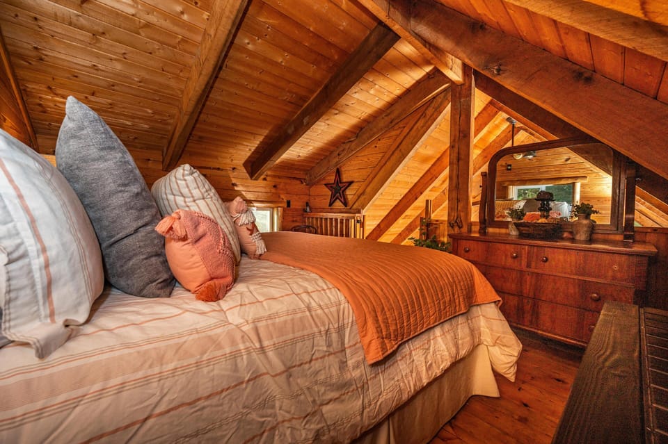 Attic bedroom with sloped pine ceilings, queen bed dressed in striped linens, and a wooden dresser with mirror.