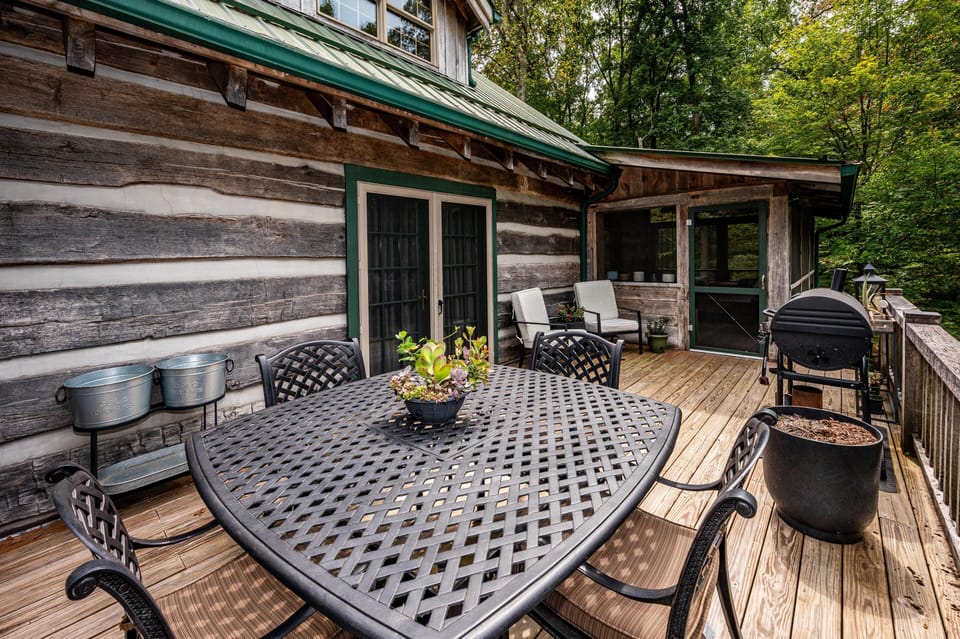 Outdoor deck with black lattice metal dining set, two cushioned armchairs, and charcoal grill surrounded by forest.