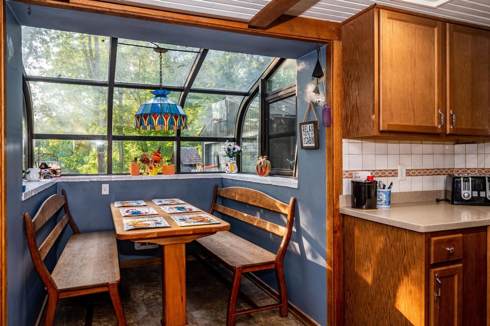 Breakfast nook with wood benches and table under a stained-glass pendant, framed by curved glass windows.