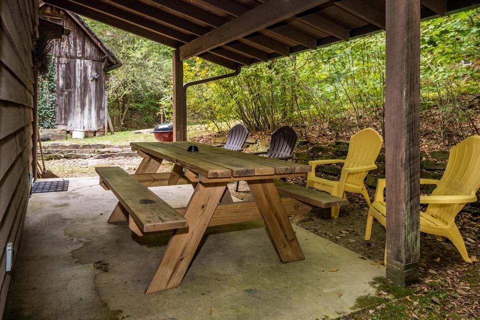 Covered patio with a wooden picnic table, yellow and brown Adirondack chairs, and charcoal grill nearby.