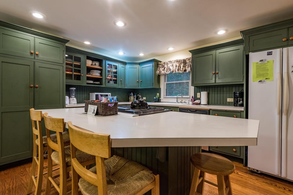 Green cabinetry surrounds the kitchen with a large white island, wooden stools, and a window over the sink.