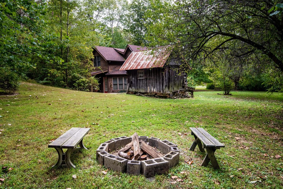 Outdoor fire pit encircled by two weathered wooden benches on a grassy yard with trees and rustic cabin.