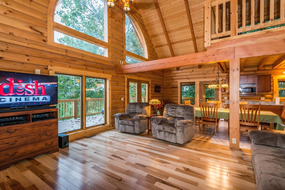 Living room and dining area with vaulted wood ceilings, plush recliners, and large windows overlooking the deck.