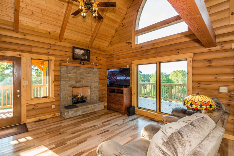 Stone fireplace with a wooden mantel flanked by log walls, a flat-screen TV on a chest, and natural light from sliding doors.