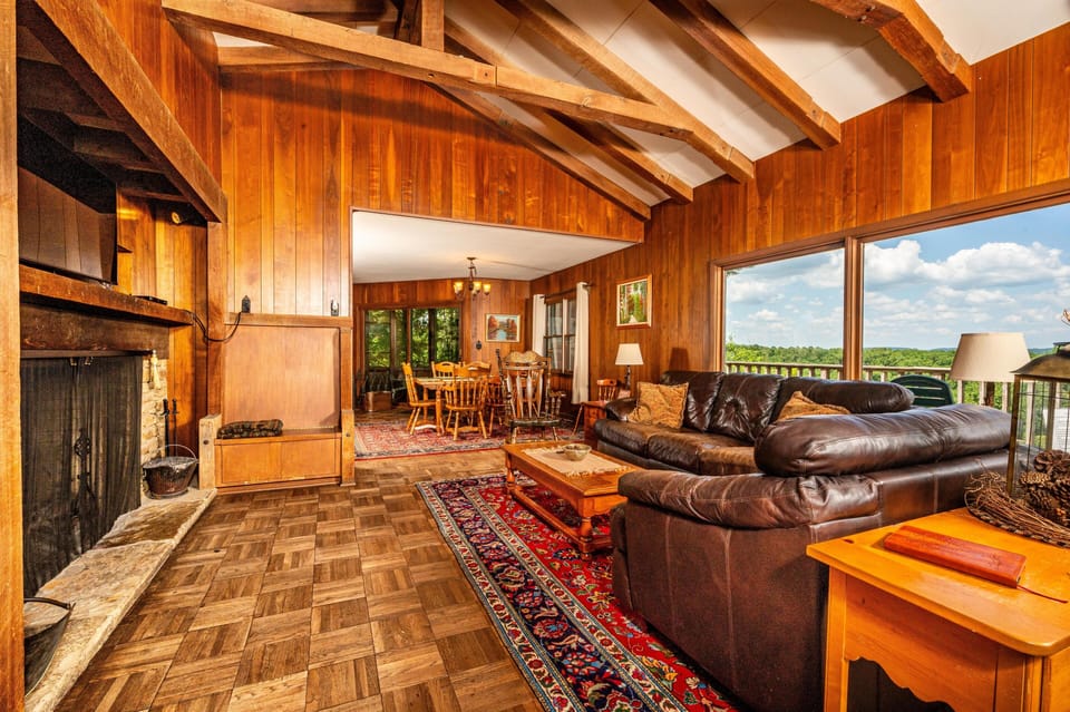 Vaulted ceiling with exposed wood beams highlights the living room’s leather sectional and large windows overlooking green hills.