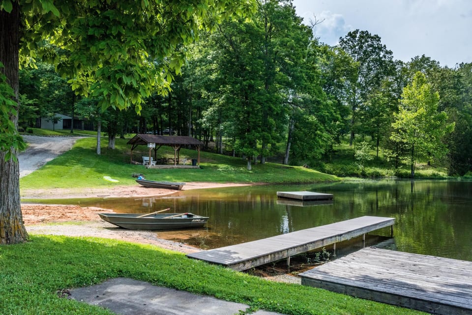 Calm lake shore with wooden docks, two rowboats, sandy beach, and a covered picnic area surrounded by trees.
