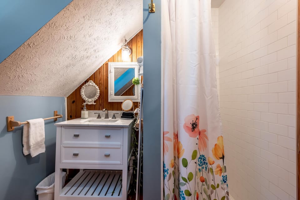 Sloped ceiling frames a white vanity with marble countertop beside a shower with floral curtain and subway tile surround.