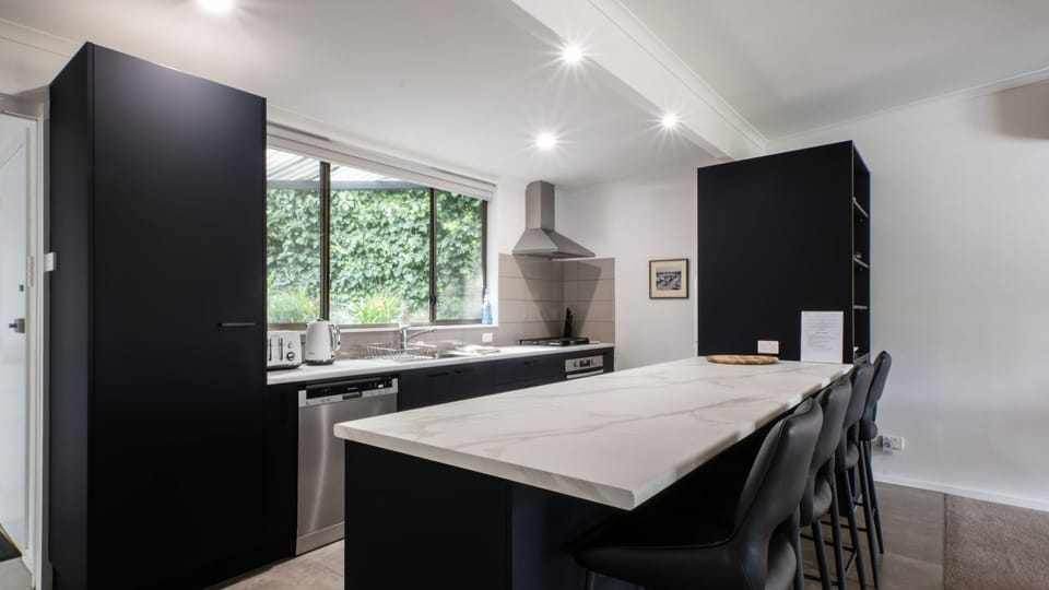 Kitchen with large breakfast bench, gas cooktop, oven, dishwasher, microwave, fridge and an abundance of cutlery, crockery, glassware, pots and pans, and a coffee plunger. 