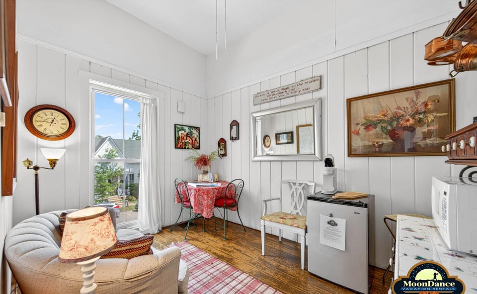 Sunlight brightens the sitting area with cream couch, red-patterned tablecloth, and rustic hardwood floors.