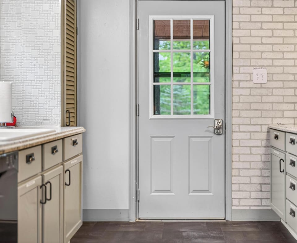 Gray door with window panels opens to a leafy outdoor view, flanked by light brick and soft-gray kitchen cabinets.