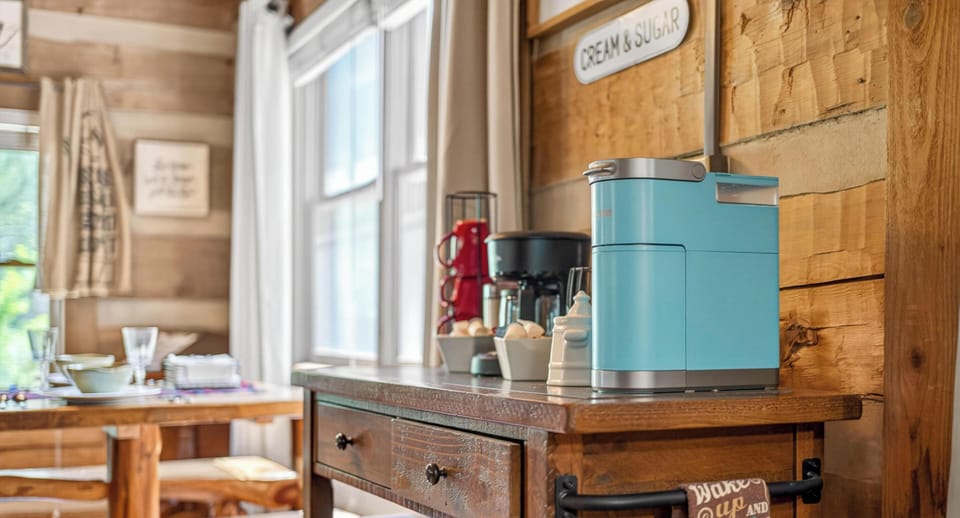 Light streams through white-trimmed windows onto a rustic wooden sideboard with a blue coffee maker and red mugs.