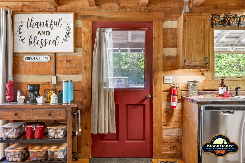 Red back door with window and beige curtain separates the kitchen’s log walls and wood cabinet.