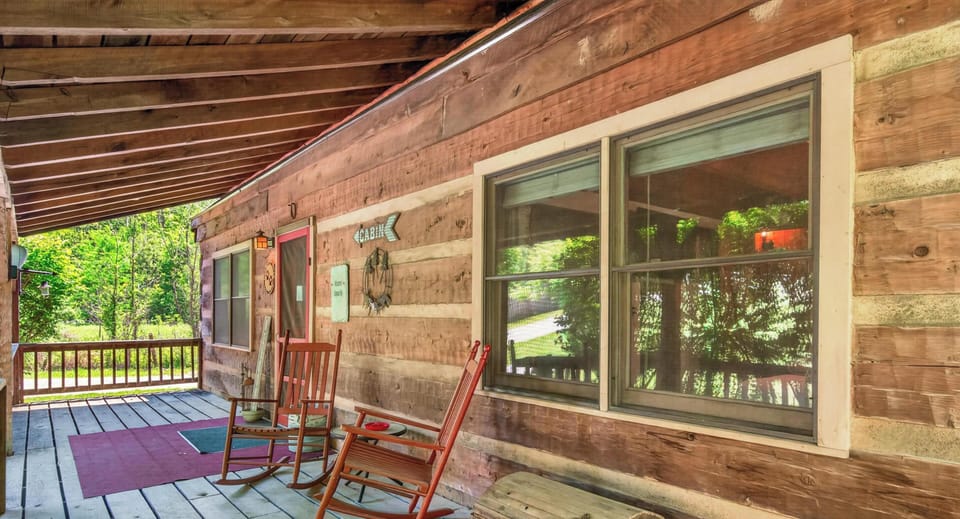 Wide wooden porch with red rocking chairs, a rustic chest, and view of green trees beyond the railing.