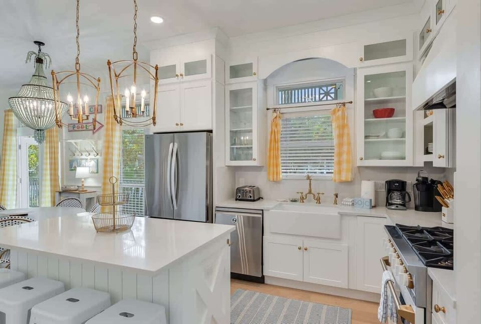 - Bright kitchen with island, brass lighting, and stool seating, ideal for casual bites or conversation - Farmhouse sink under a window with yellow curtains adds charm & coastal warmth - Stainless appliances & open shelves blend style & function