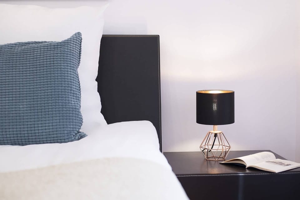 A close-up of a bedside table with a lamp, next to a bed with crisp, white bedding. The lighting creates a warm ambiance.