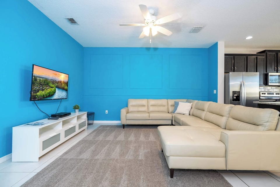 Bright living room with a bold teal accent wall, comfy leather sectional with chaise, ceiling fan, and wall‑mounted smart TV-perfect for cozy movie nights in a modern coastal vibe.