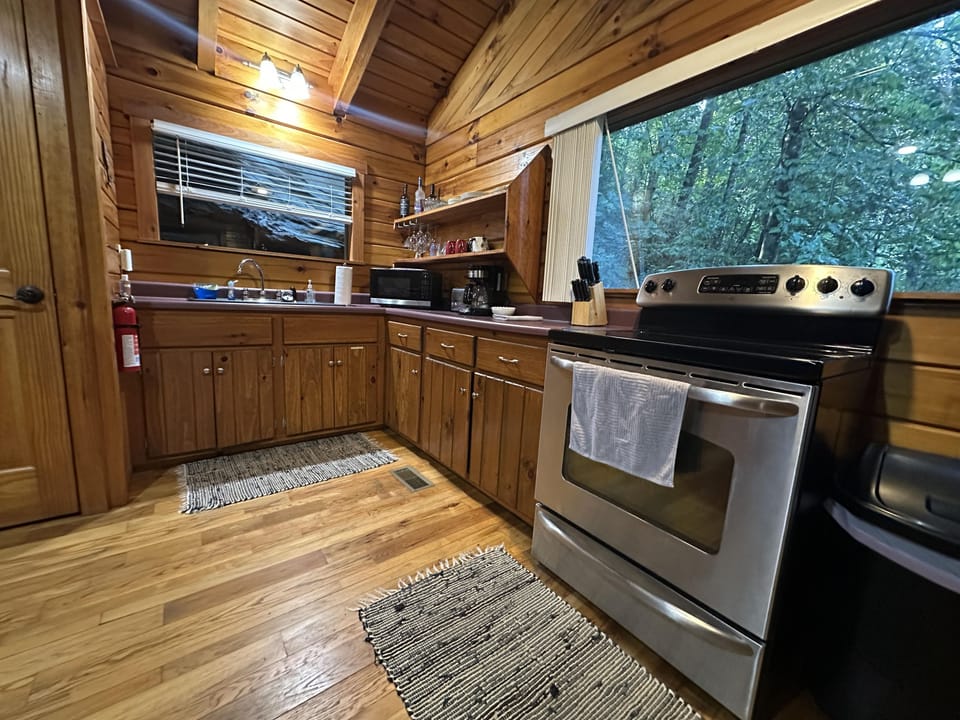 Kitchen with stainless steel stove, wooden cabinetry, dual rugs on hardwood floor, and large window overlooking forest.