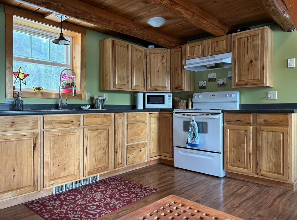 Kitchen features natural wood cabinets, black countertops, white stove, microwave, and a window with a star-shaped stained glass.