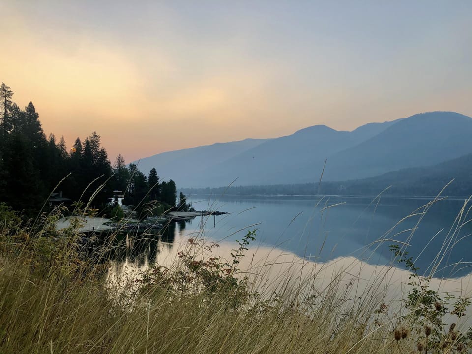 Tall dry grasses frame a calm lake reflecting blue mountains under a hazy sunset sky.