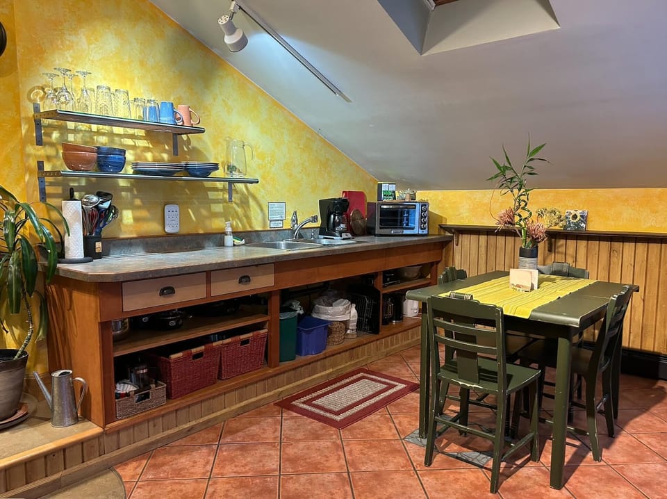 Yellow textured walls frame an angled ceiling above open kitchen shelves and a green dining table with six chairs.