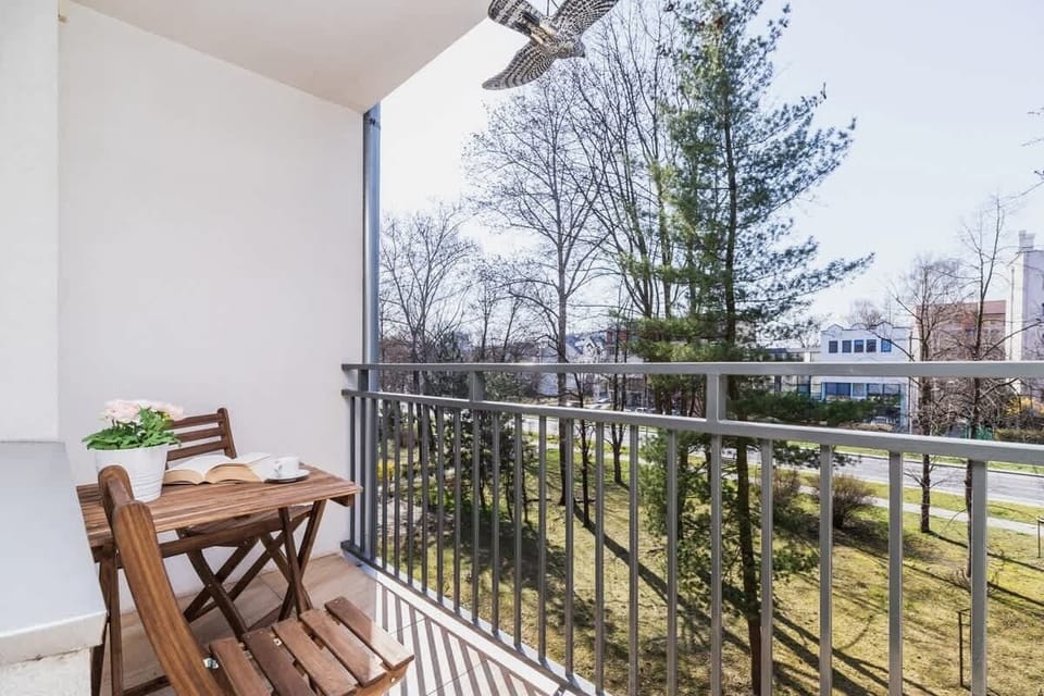A balcony with outdoor seating, featuring a small table and chairs. The view shows a green outdoor area with trees and neighboring buildings.