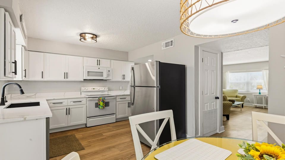 Glass-top dining table with sunflower centerpiece overlooks white kitchen cabinetry and stainless steel refrigerator.