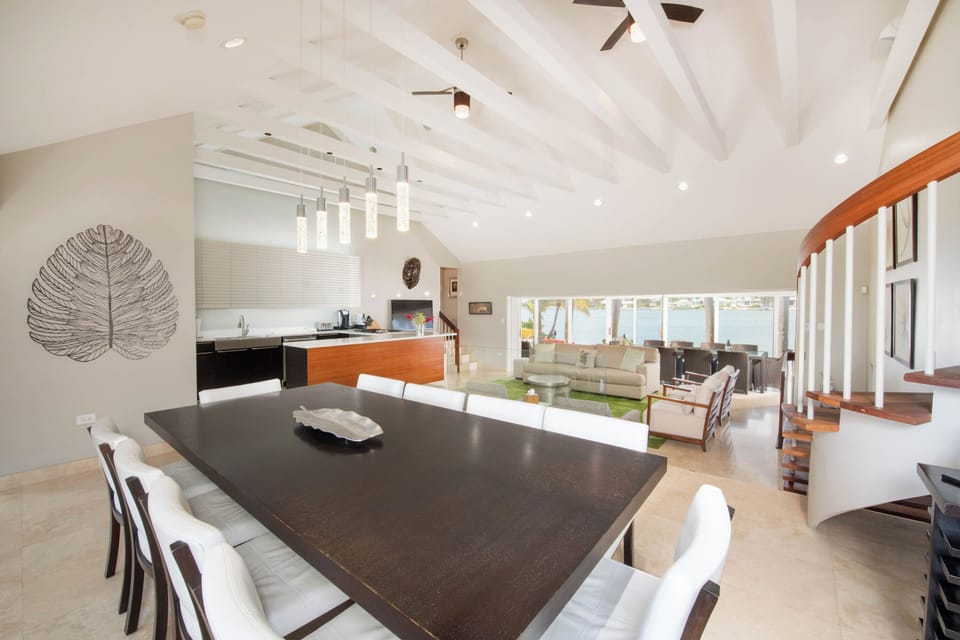 The main villa's great room features vaulted ceilings, an open concept design with multi-levels, and waterfront views throughout. The formal dining table seats 10 guests.