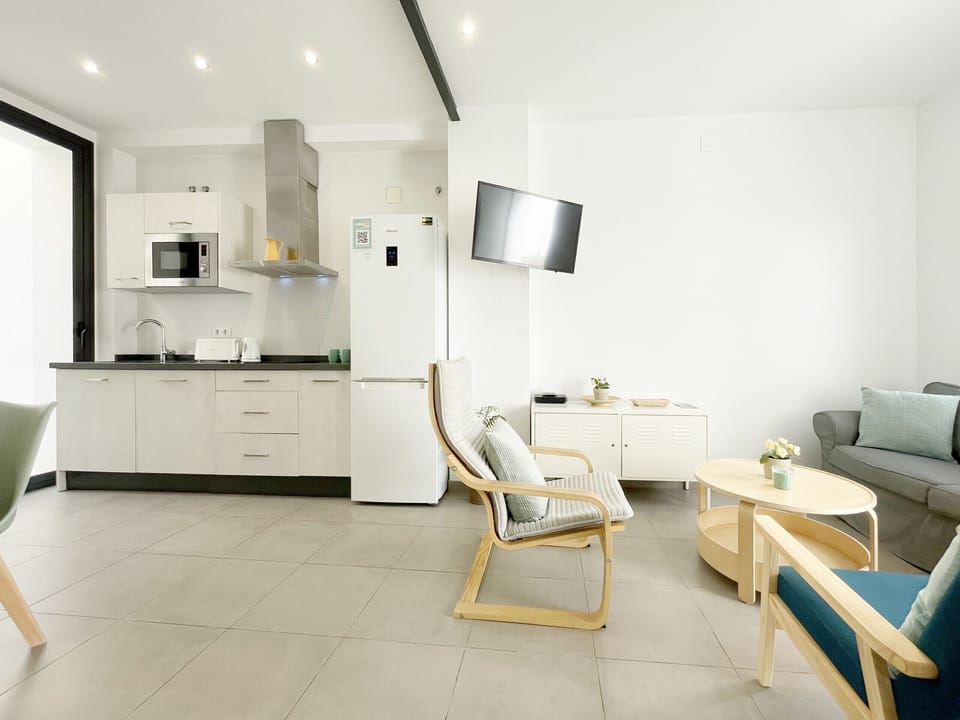 Open-plan living room and kitchen with a modern and bright design. White kitchen on the left and living area with wooden chairs and a gray sofa.