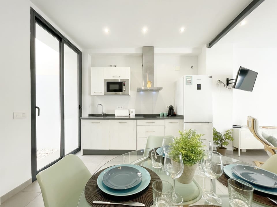Open-plan living room and kitchen with a modern and bright design. White kitchen on the left and living area with wooden chairs and a gray sofa.