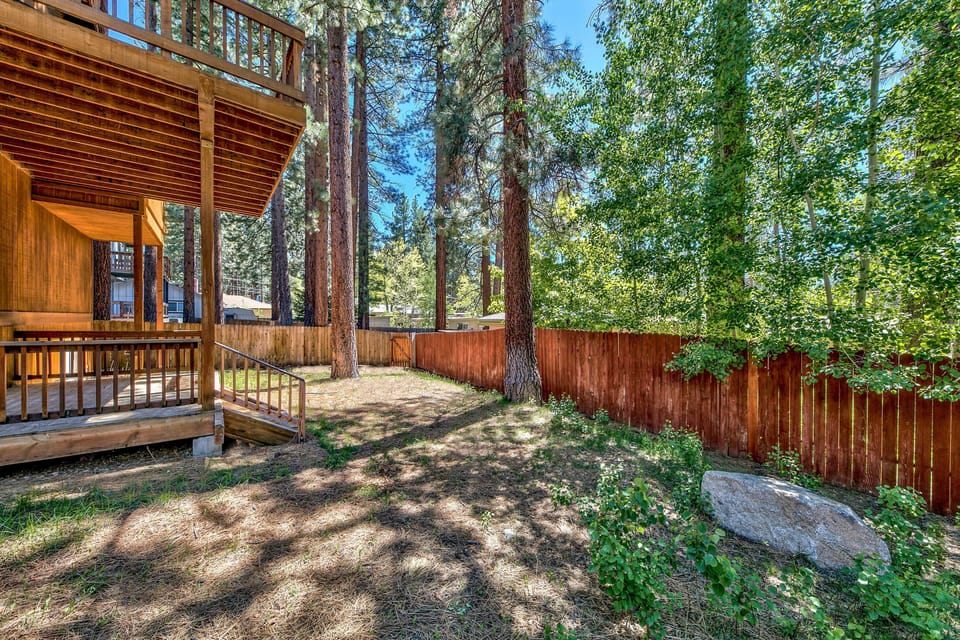 Fenced backyard with tall pine trees, shaded grassy area, wooden deck, and a large rock near the fence.
