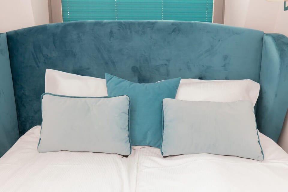 A serene bedroom featuring a bed with white linens, turquoise pillows, and a bold turquoise headboard. The window adds brightness to the room.