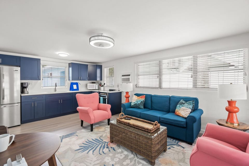 Siesta Key coastal living room featuring a teal sofa, pink accent chairs, and blue kitchen backdrop—crafted for breezy comfort and effortless seaside style.