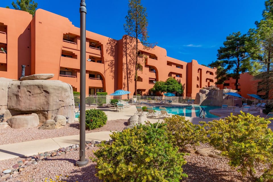 Outdoor pool area with rock waterfall features, blue umbrellas, lounge chairs, and desert landscaping.