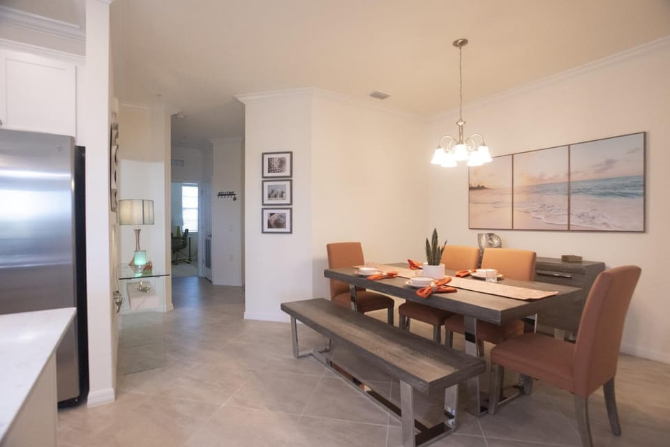 Rectangular dining table with wood bench and rust-colored chairs under a five-light chandelier and beach triptych.