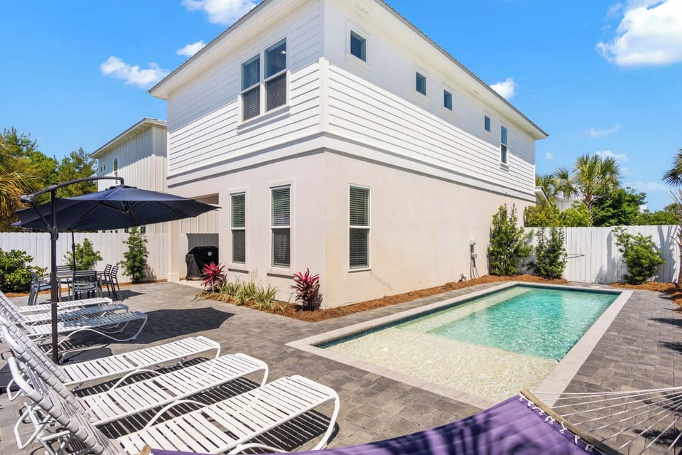 Unwind by your private pool surrounded by brick paving, sun loungers under a large navy umbrella, and a cozy hammock near lush white fencing.