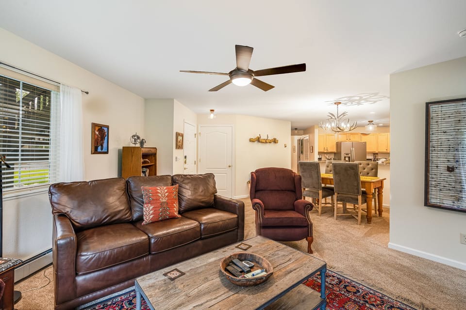 A living room with a brown leather sofa, red armchair, wooden coffee table, and ceiling fan. The room leads into a dining area with a table and chairs and a kitchen with wooden cabinets.