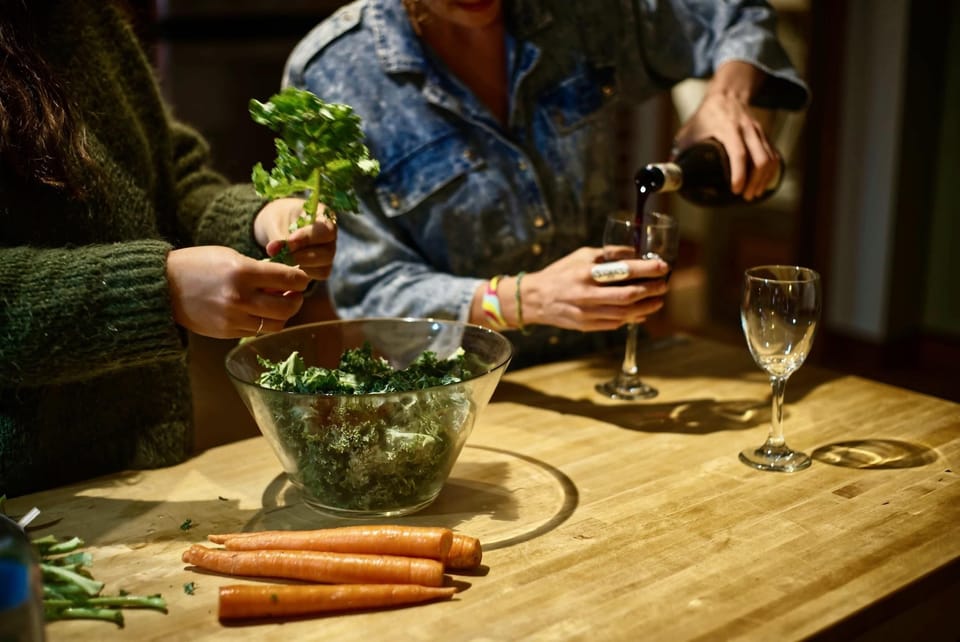 Hands tear kale into a glass bowl on a natural wood kitchen island as red wine is poured into glasses.