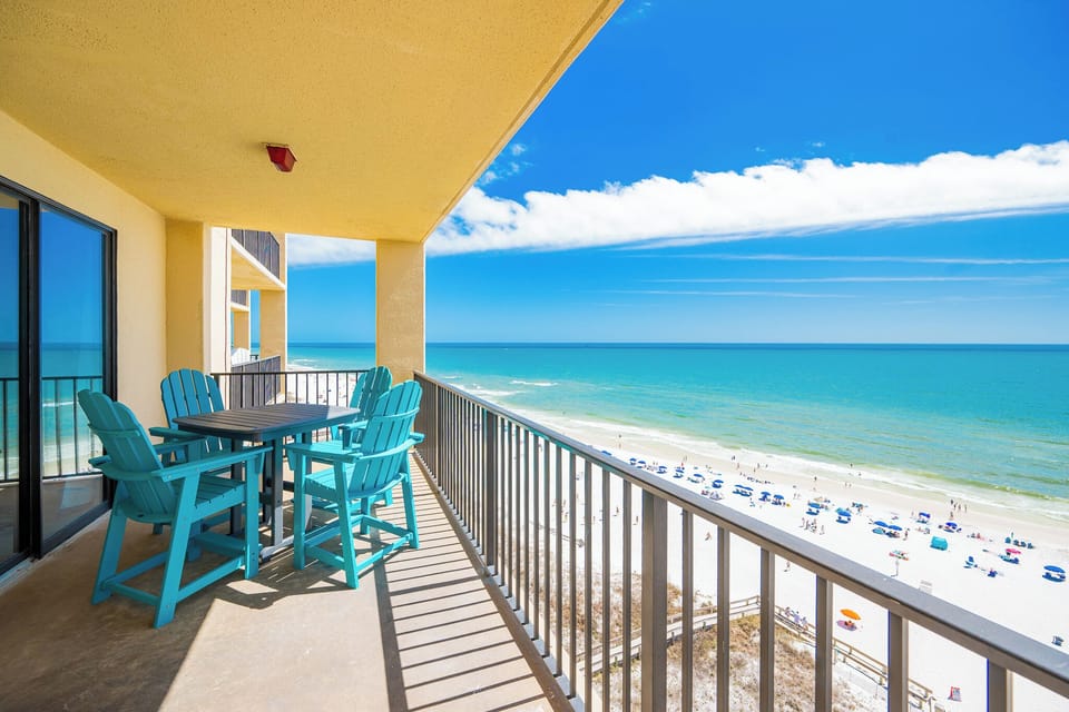 The balcony features vibrant turquoise seating and a table, offering a panoramic view of the beach and sparkling waters
