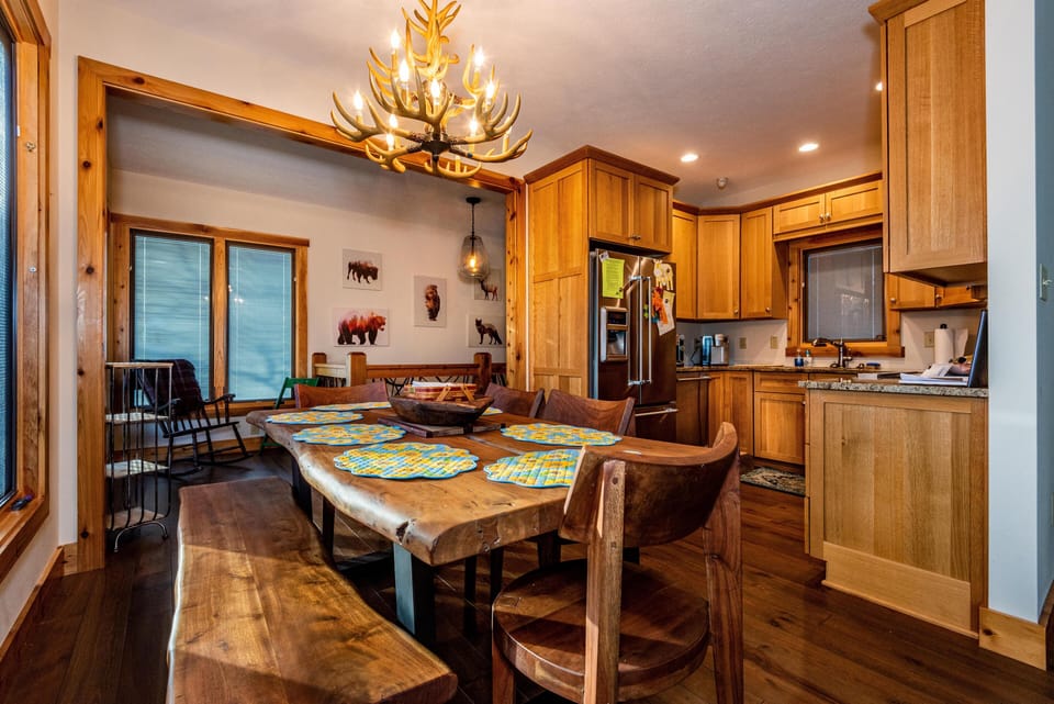 Live-edge wood dining table with quilted placemats sits beneath an antler chandelier by a windowed nook.
