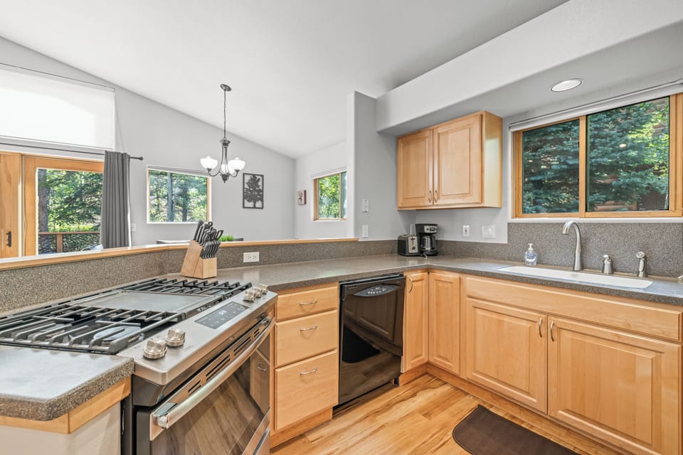 Contemporary kitchen featuring light wood cabinetry, sleek appliances, and a gas cooktop. Large windows fill the space with natural light while offering serene views of the surrounding forest. Perfect for preparing meals after a day of mountain adventures.