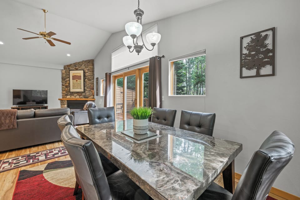 Stylish dining room featuring a polished stone-top table and plush seating for six. Large windows and glass doors frame a peaceful forest backdrop, seamlessly connecting to the cozy living area and stone fireplace.