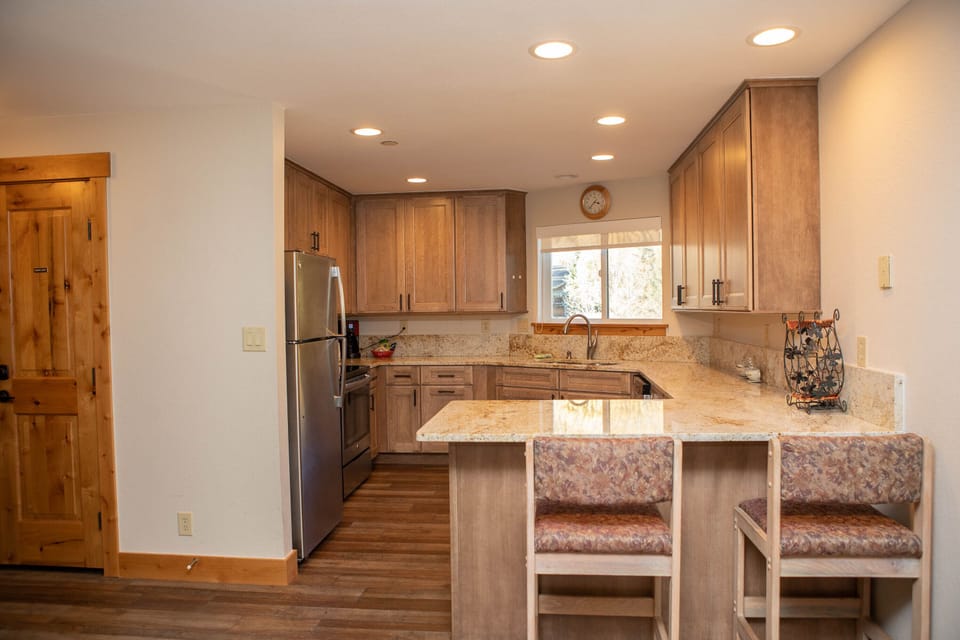 A modern kitchen with wooden cabinets, granite countertops, stainless steel appliances, a small window above the sink, and a breakfast bar with two padded stools.