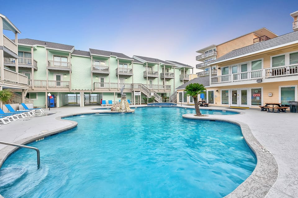 Spacious resort-style pool with tropical fountain centerpiece, surrounded by colorful multi-level buildings and palm trees under clear skies.