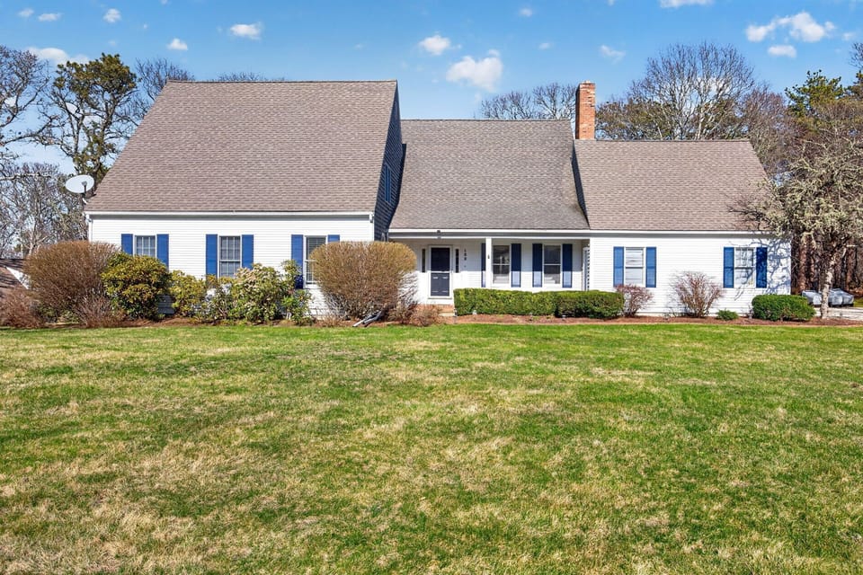 Charming white Cape Cod home with blue shutters sits on spacious green lawn surrounded by mature trees.