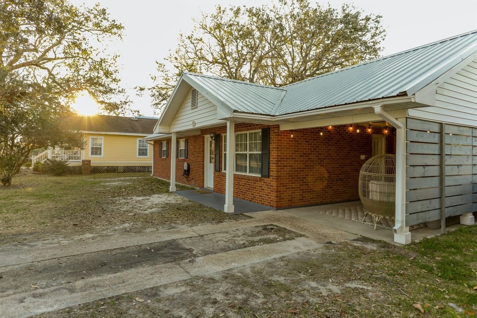 Cozy brick home with a metal roof and string lit porch welcomes the golden glow of sunset in a peaceful neighborhood setting.