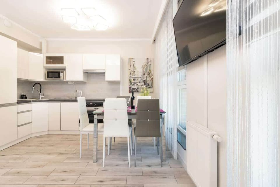  A kitchen with white cabinets, a small dining table with chairs, and neutral decor. The layout is practical, and large windows with blinds bring in natural light.

